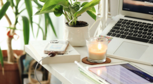 Close up of white desk with macbook, ipad, iphone, and small candle on top. Large, green, potted, indoor plants are in the background nearby.